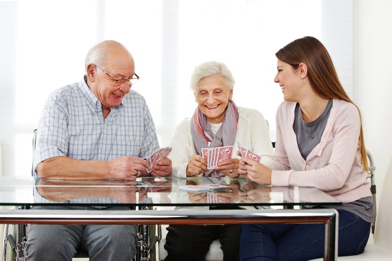 Junge Frau spielt mit zwei glücklichen Senioren zusammen Karten im Pflegeheim